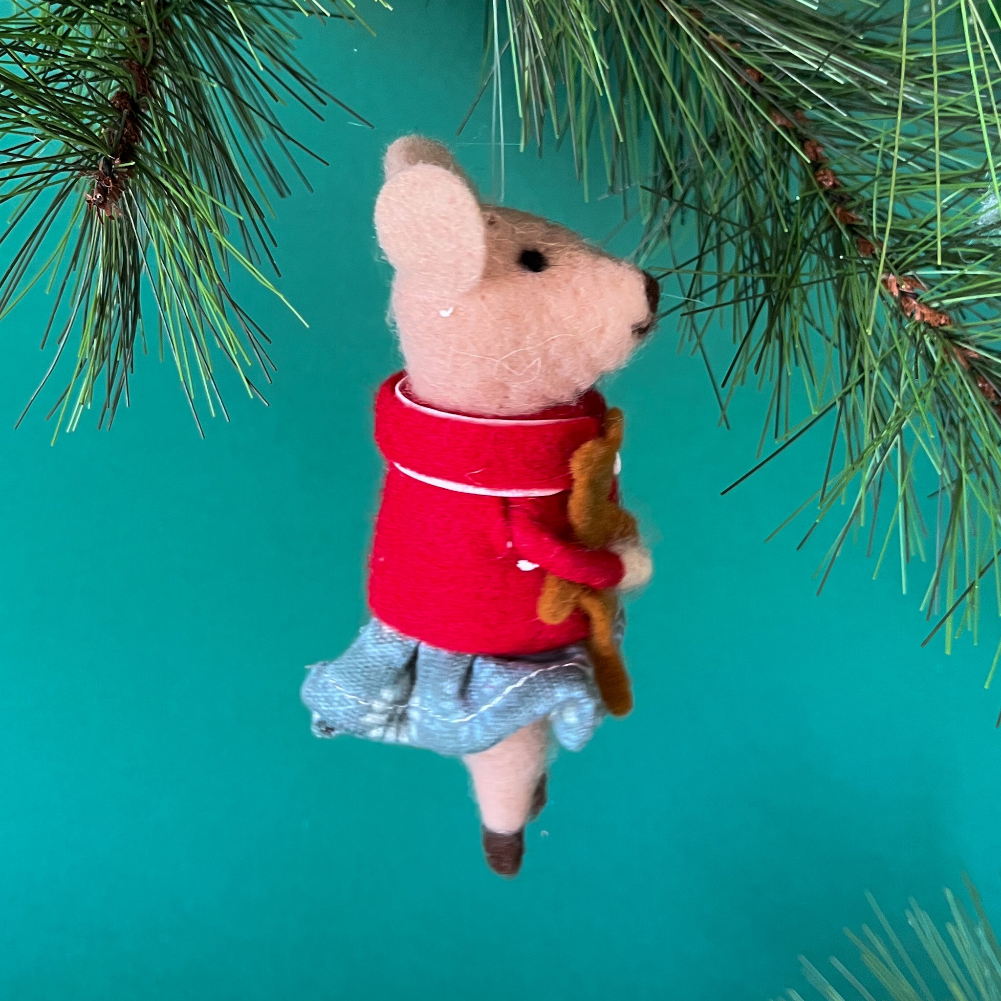 side of a felt mouse ornament wearing a red jacket and gray skirt holds a small brown teddy bear, hanging from a pine branch against a teal background.