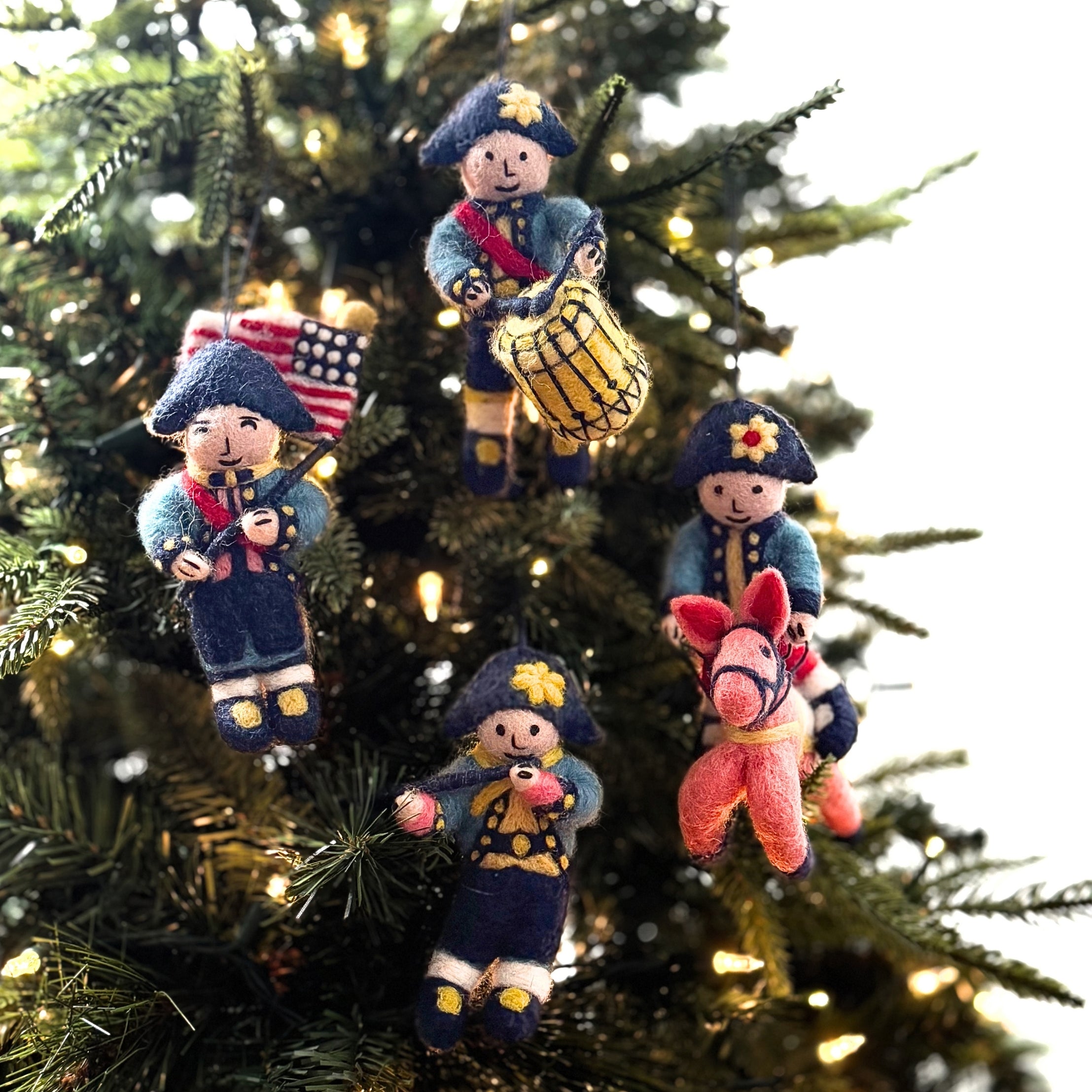 Four felt colonial soldier patriotic Christmas ornaments hanging on a lit Christmas tree. The ornaments feature different soldier designs: one with a flag, one with a drum, one on horseback, and one with a rifle.