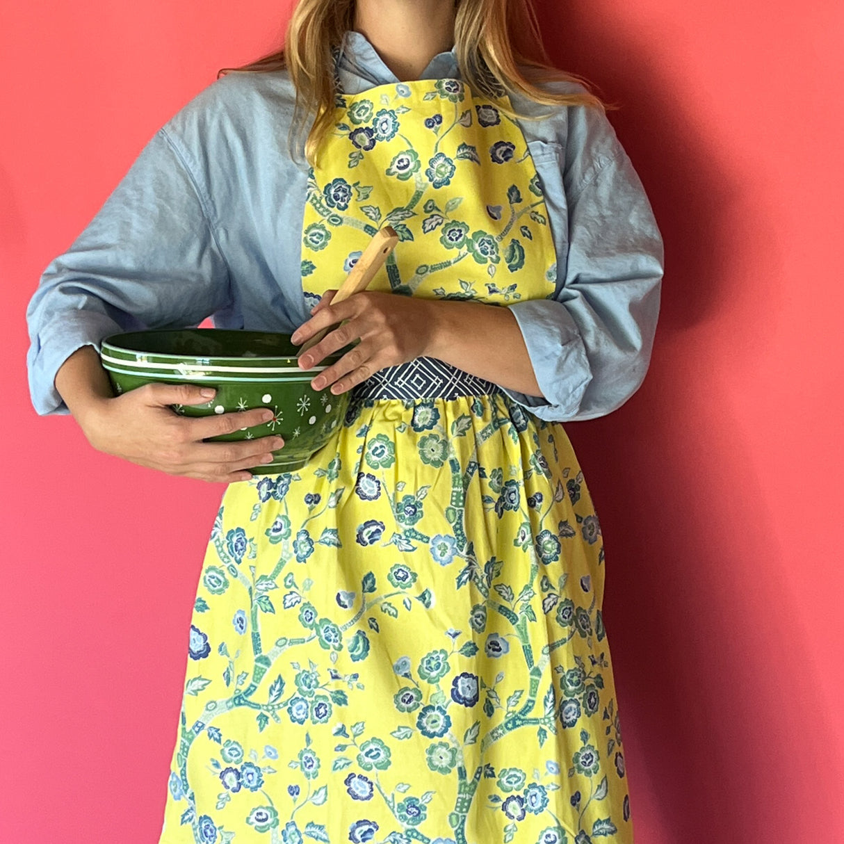 Person wearing a Madcap cottage yellow floral apron holding a green bowl against a red background