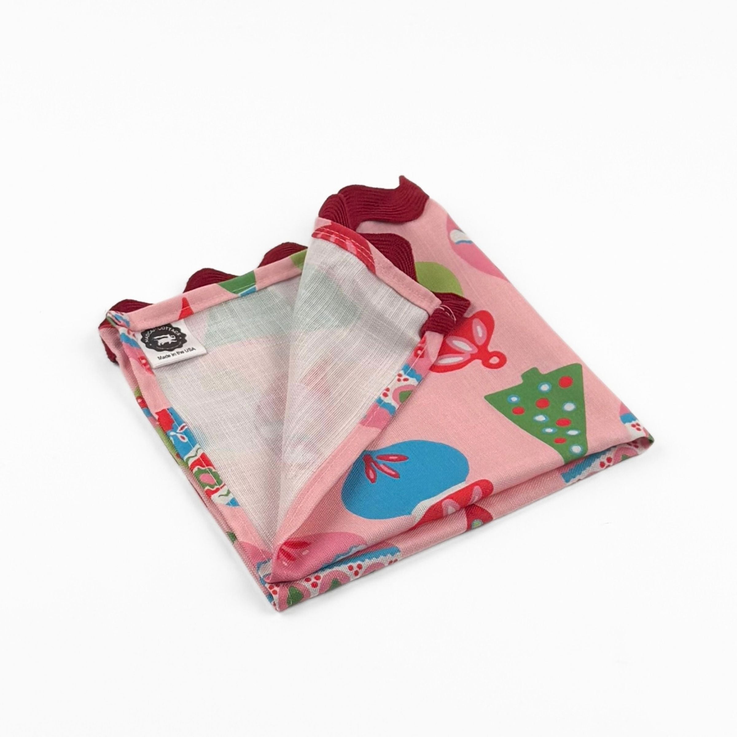 Folded Pink Christmas dinner napkins featuring colorful ornaments, festive trees, and red scalloped trim for tablescapes.