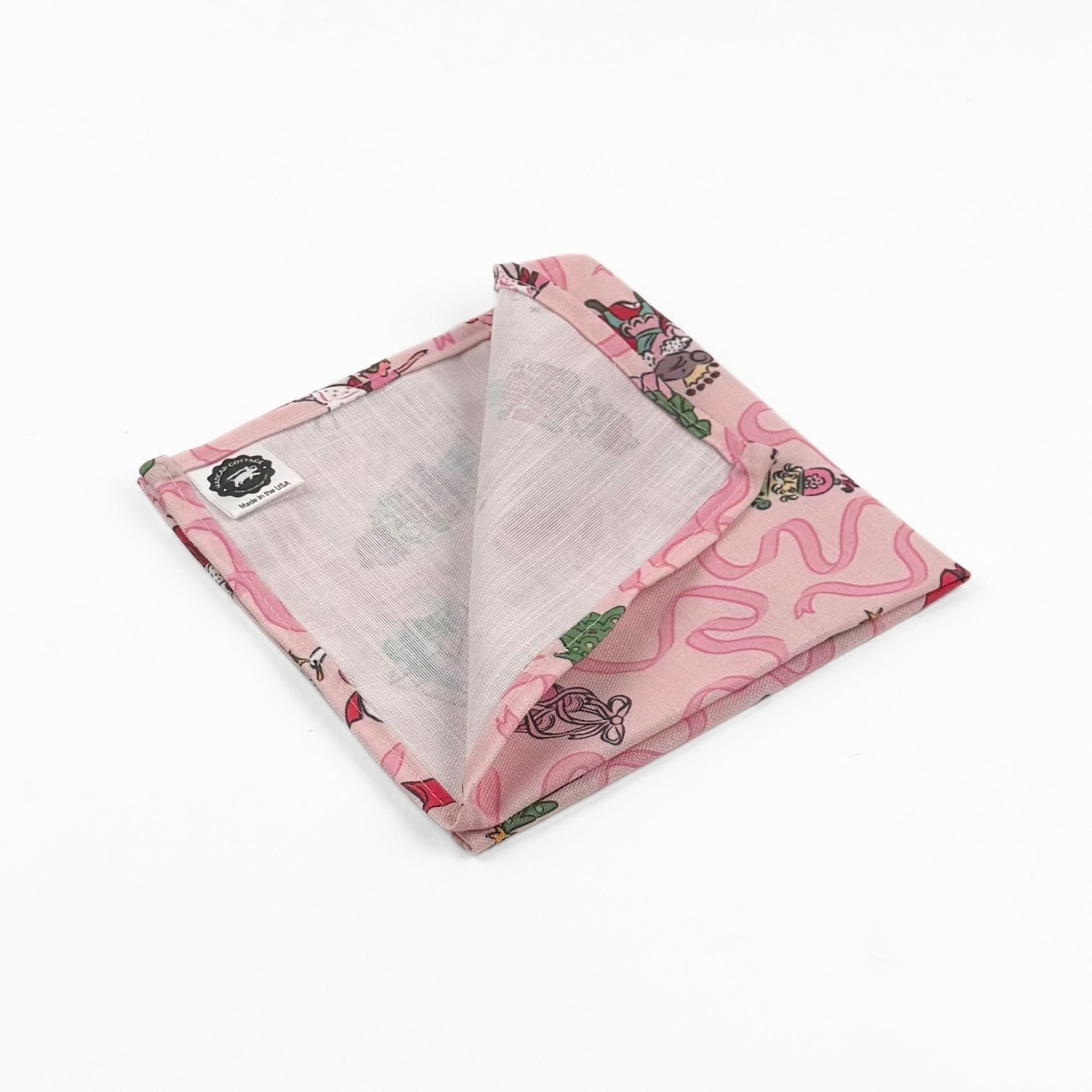 Folded Pink Christmas dinner napkins featuring swirling ribbons, nutcrackers, ballerinas, festive gifts, and whimsical holiday illustrations.
