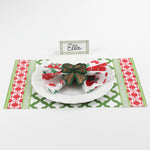 Christmas table setting featuring Bamboo Trellis Paper Place Card in forest green, white plate with floral napkin and woven bow napkin ring on red and green patterned placemat.