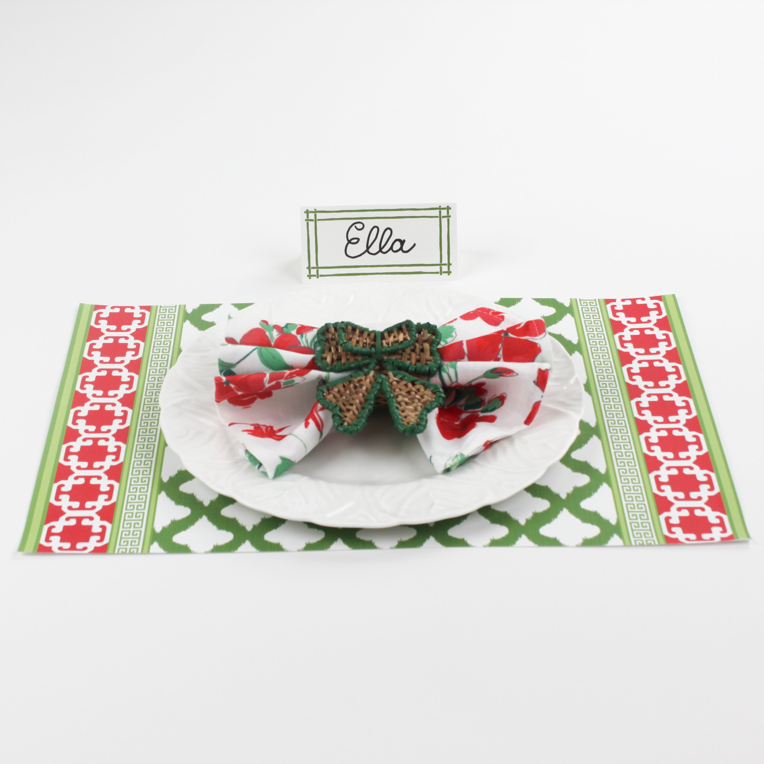 Christmas table setting featuring Bamboo Trellis Paper Place Card in forest green, white plate with floral napkin and woven bow napkin ring on red and green patterned placemat.