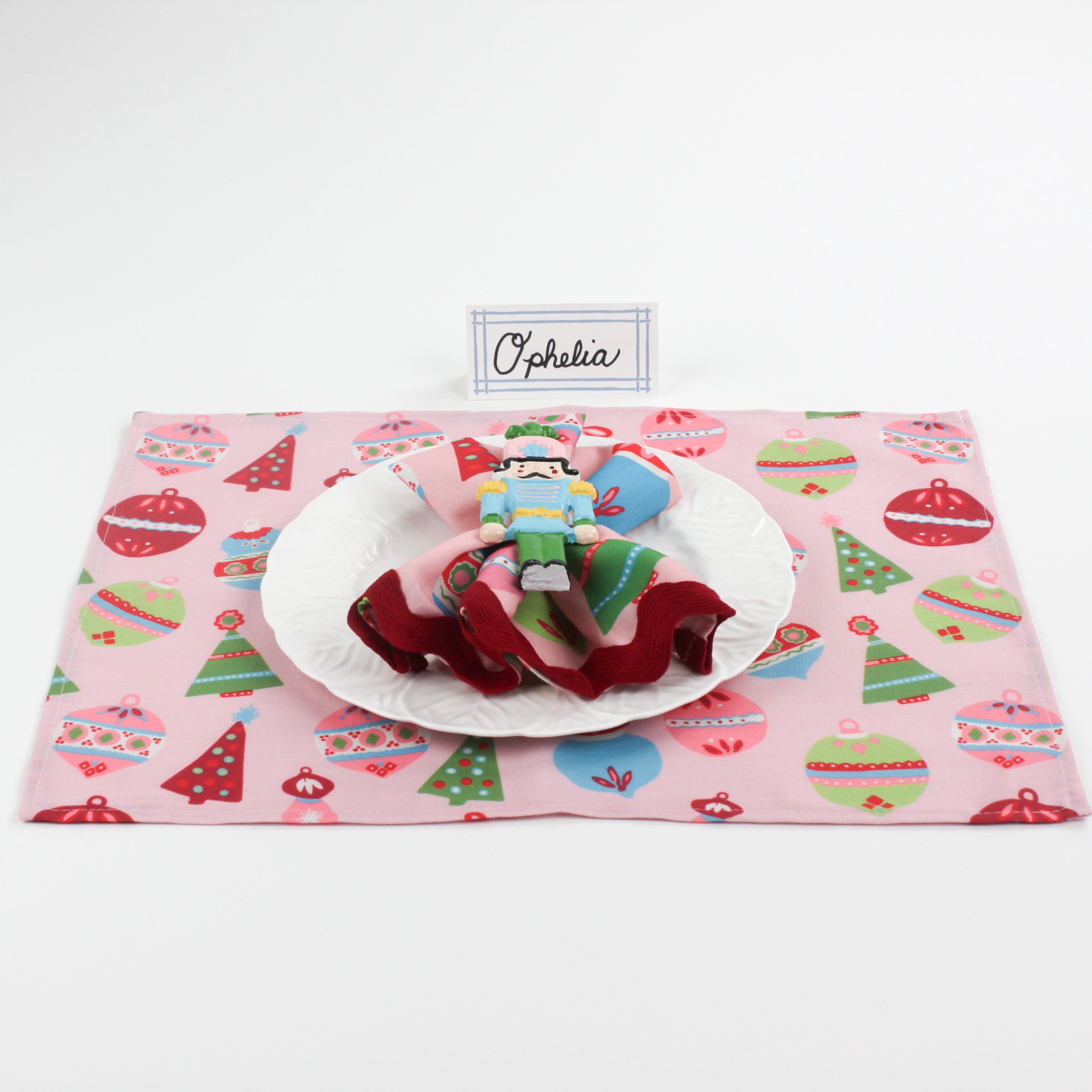 Christmas table setup featuring Bamboo Trellis Paper Place Card in pool blue, white plate, nutcracker napkin, and pink holiday placemat with ornaments and trees.