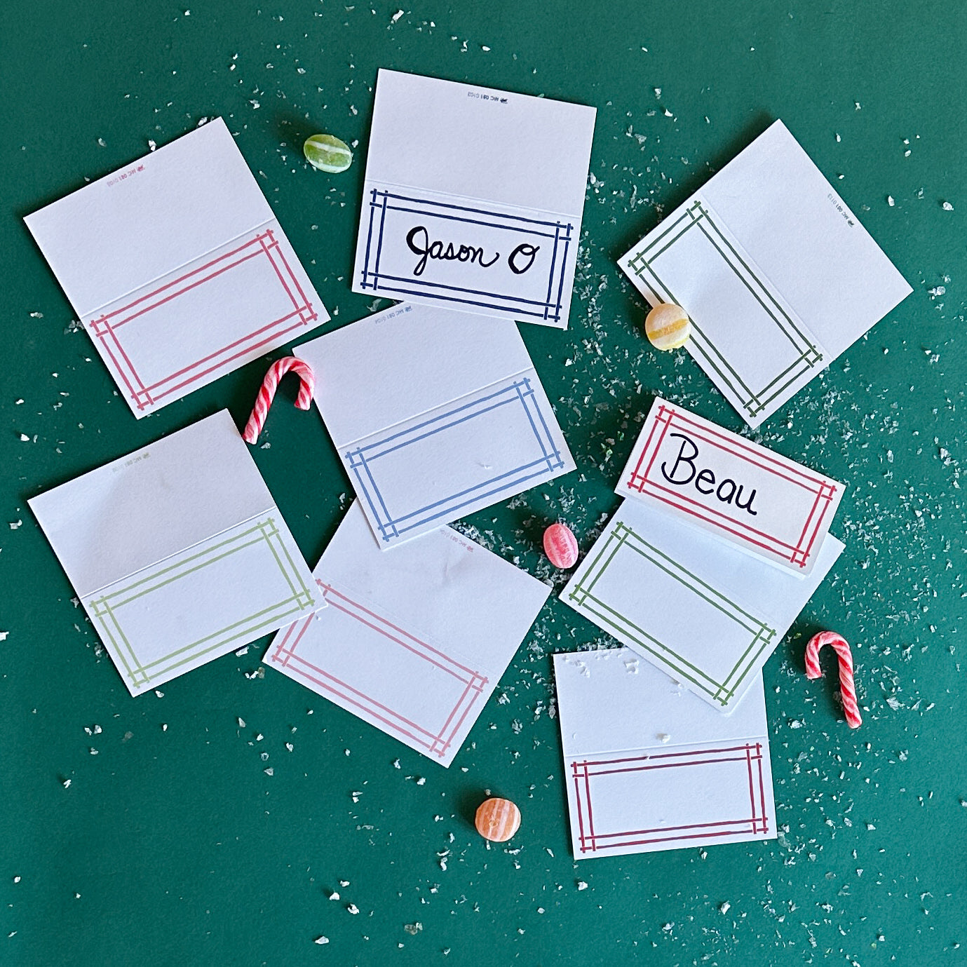 Assorted Bamboo Trellis Paper Place Cards with various colors and text, displayed on a green background with candy cane pieces scattered around.