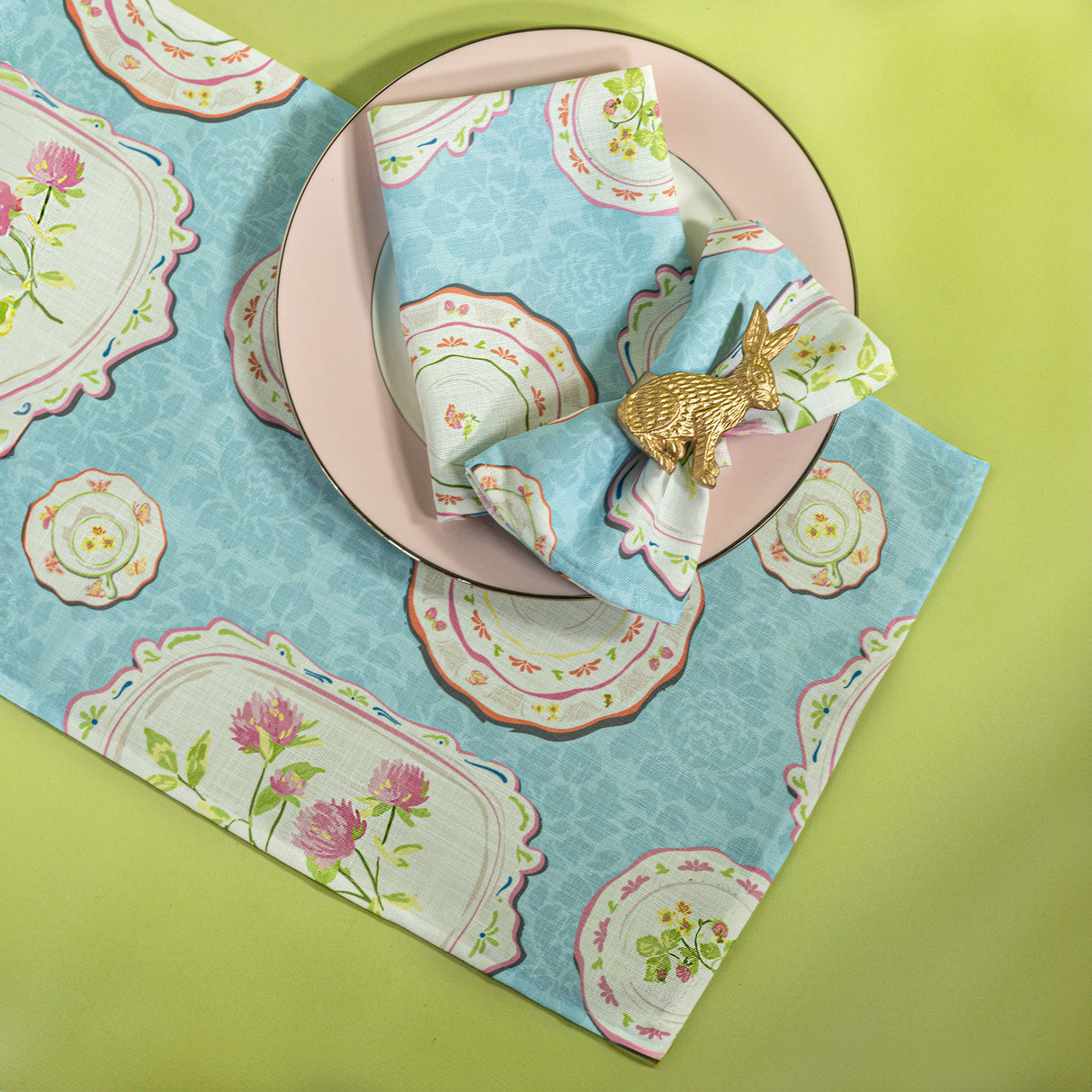 blue society hill vintage plates cloth table runner and napkins with a pink plate and bunny napkin rings on a green background.