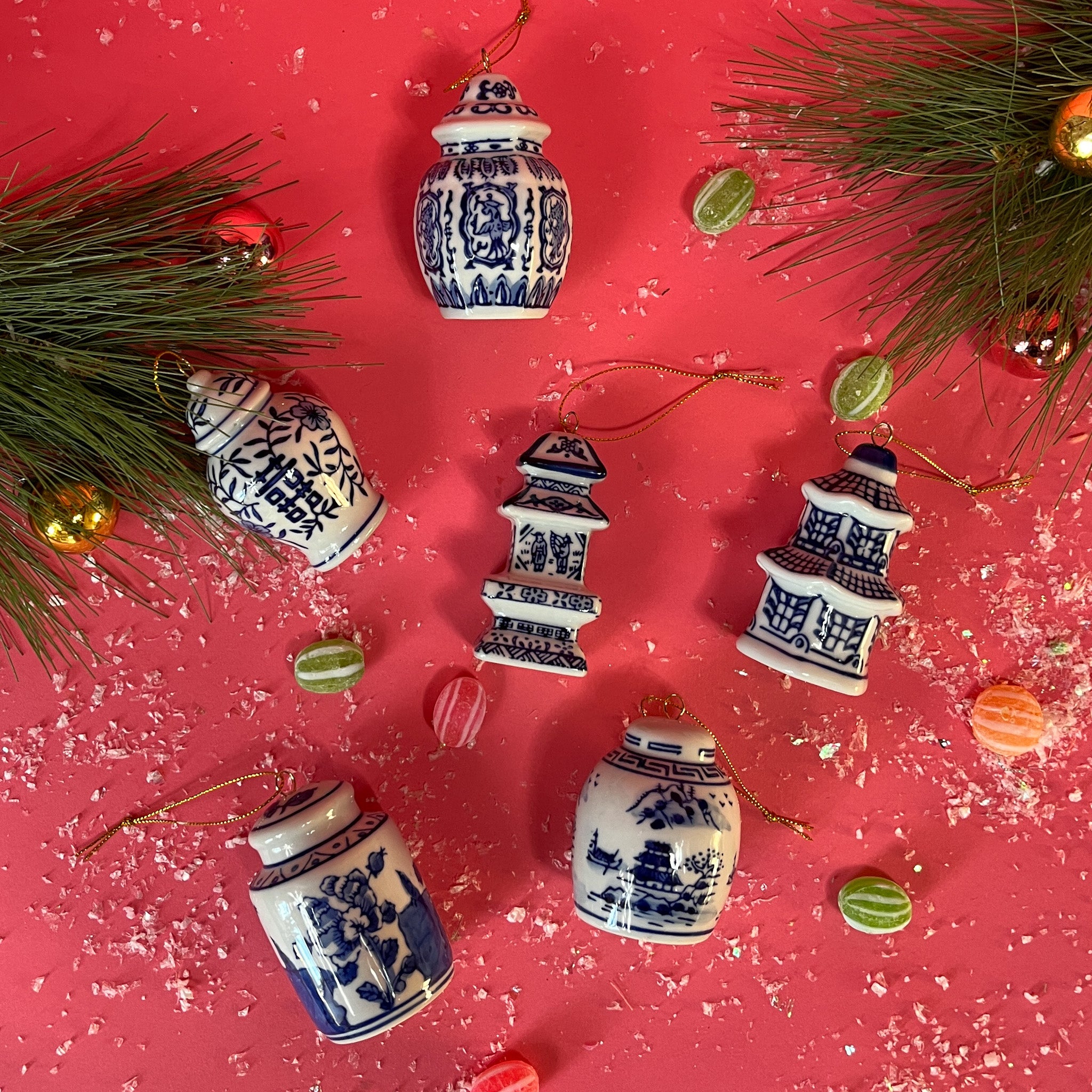 A set of six blue and white miniature ceramic ginger jars with Christmas ornaments, displayed against a red background with green pine branches and holiday decorations.