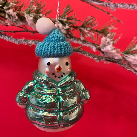 A glass christmas snowman ornament wearing a blue hat and coat against a red background