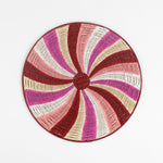 A round rattan christmas placemat with pink, red, white and yellow candy-cane like swirls. 