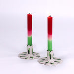 Two lit red-to-green gradient taper candles in white ceramic scalloped candle holders with green brushstroke design, displayed on a light background.