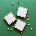 Colorful square notepads with decorative borders, displayed on a green background surrounded by candy canes and decorative balls.