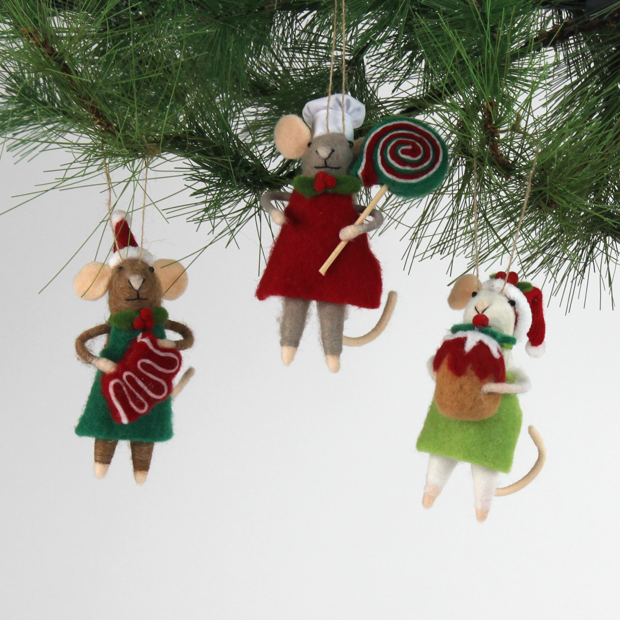 Three festive felt mouse ornaments wear holiday outfits—one holds a gingerbread cookie, another a giant lollipop, and the third a Christmas pudding. all three hanging in a pine branch