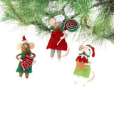 Three festive felt mouse ornaments wear holiday outfits—one holds a gingerbread cookie, another a giant lollipop, and the third a Christmas pudding. all three hanging in a pine branch