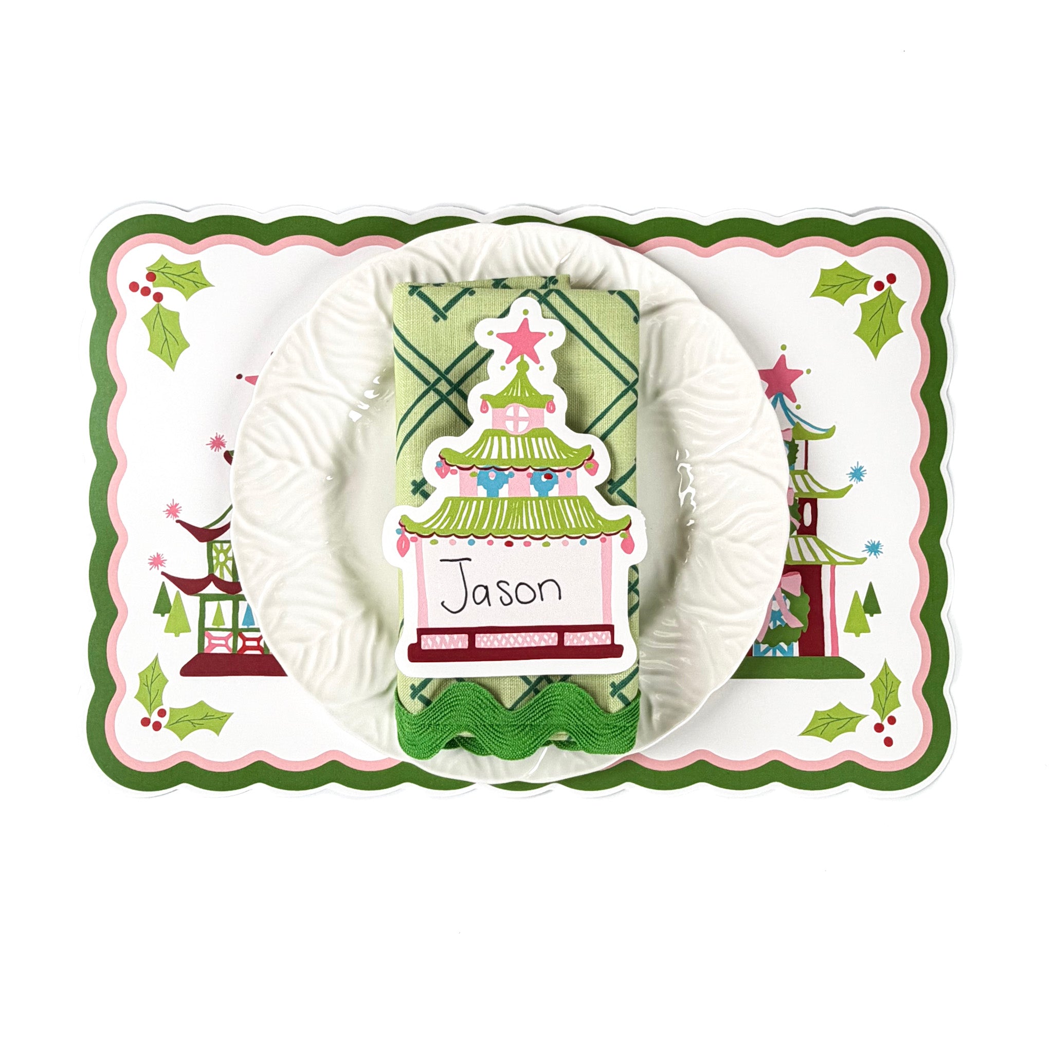 Holiday table setting featuring a Christmas pagoda decorative placemat, white ruffled plate, green patterned napkin, and pagoda-shaped paper place card with handwritten name, arranged on a white background.