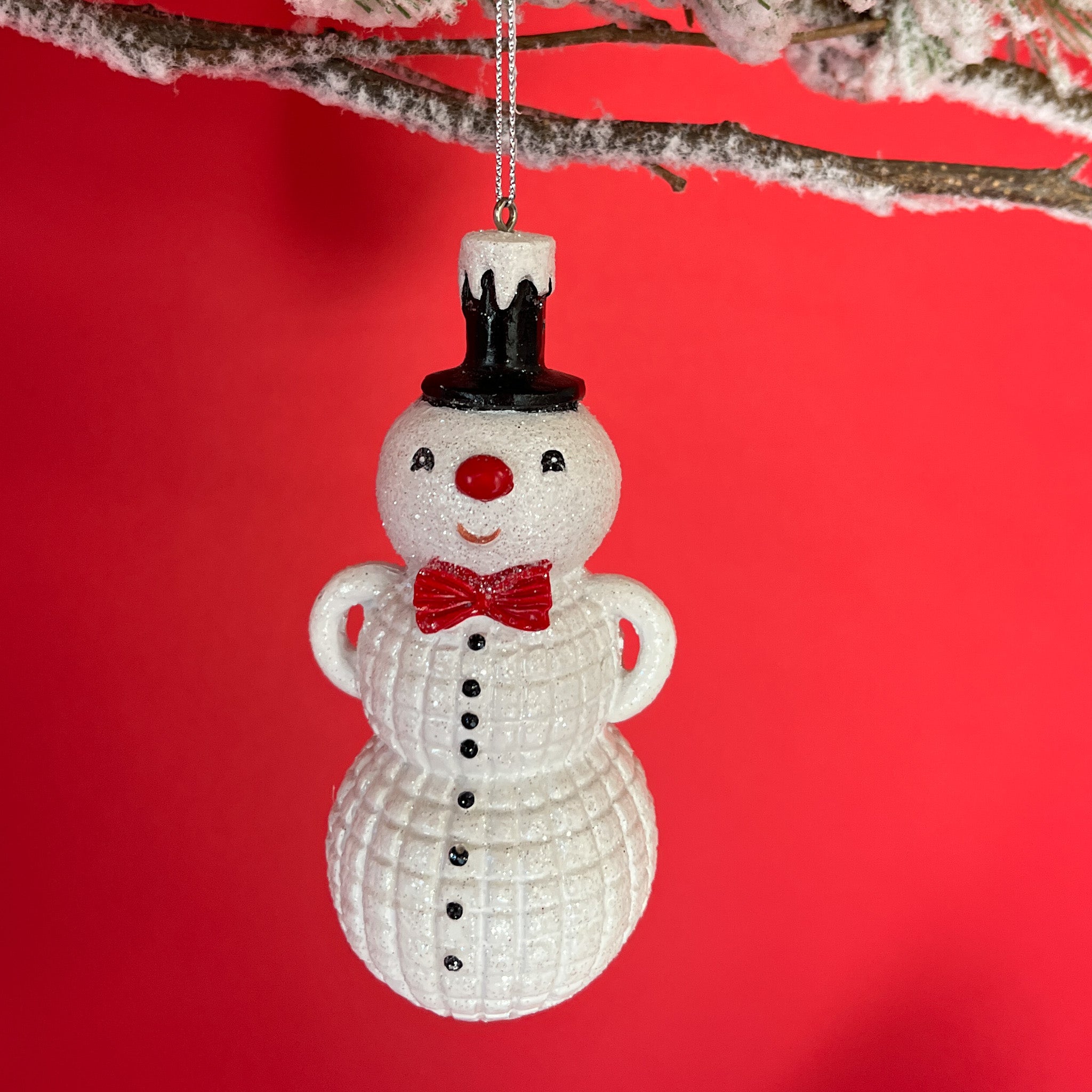 Christmas snowman ornament wearing a black top hat and red bow tie against a red background.