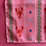 A close-up view of a cloth dinner napkin printed with lobsters,  blue bows and green stripes on a pink background