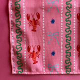 A close-up view of a cloth dinner napkin printed with lobsters,  blue bows and green stripes on a pink background