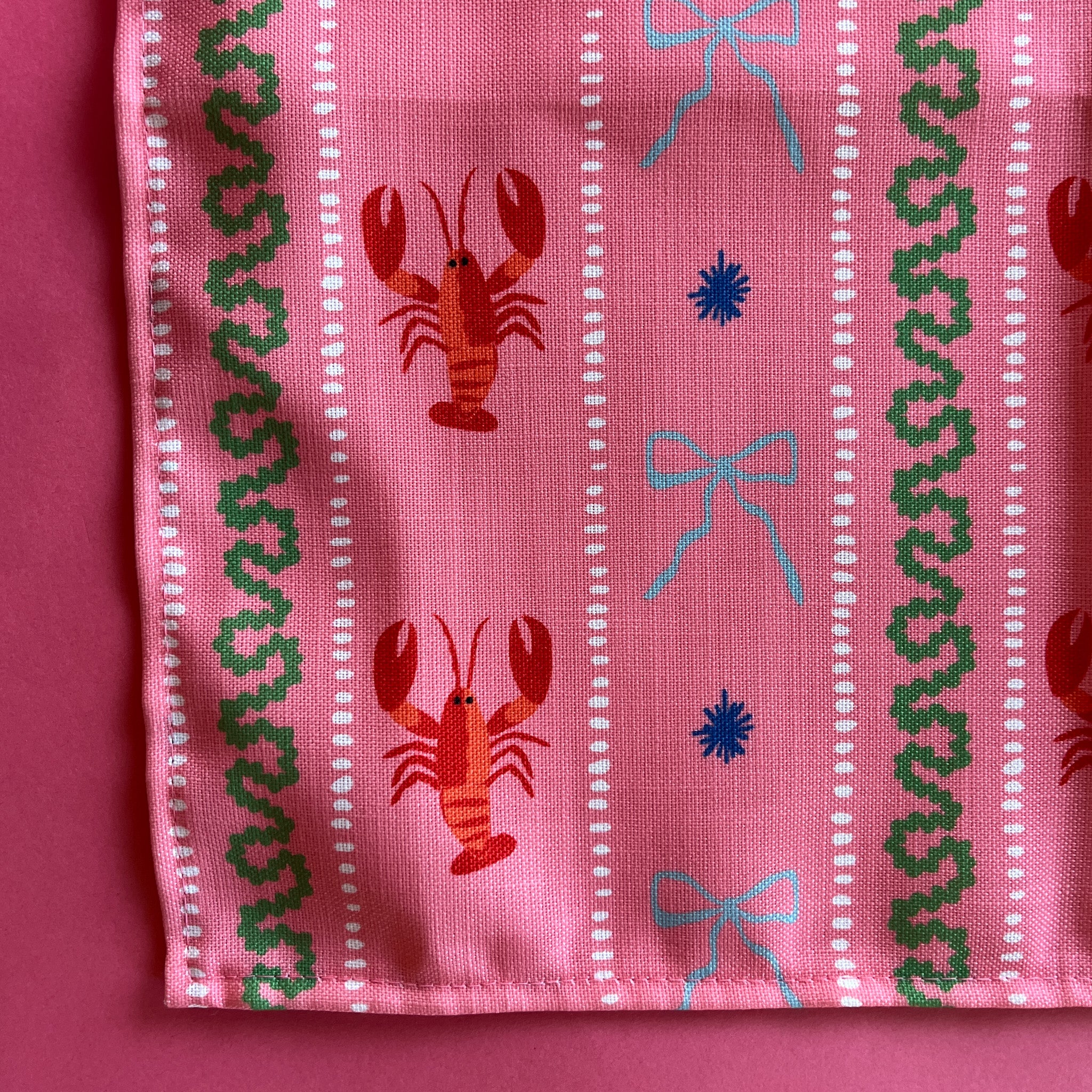 A close-up view of a cloth dinner napkin printed with lobsters,  blue bows and green stripes on a pink background