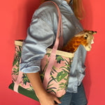 Close-up side view of a model holding the Jungle Road large canvas tote in pink and green with a toy giraffe sticking out of the tote.
