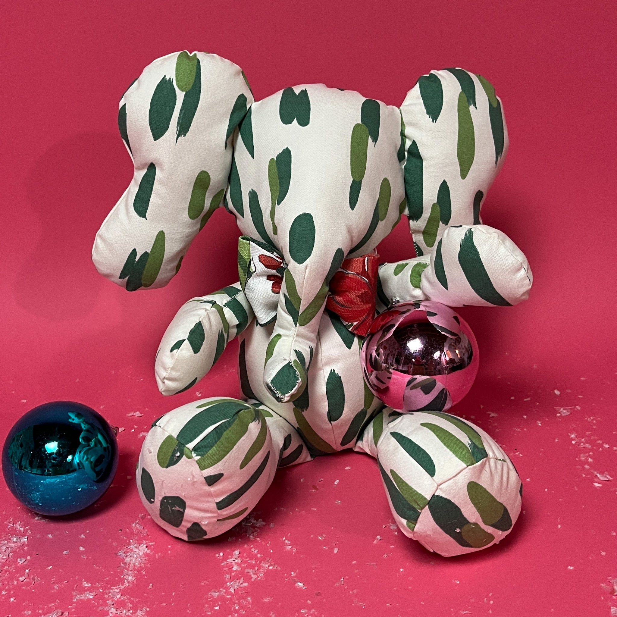 White stuffed elephant toy with green brushstroke pattern and red bow posing with colorful christmas ball.