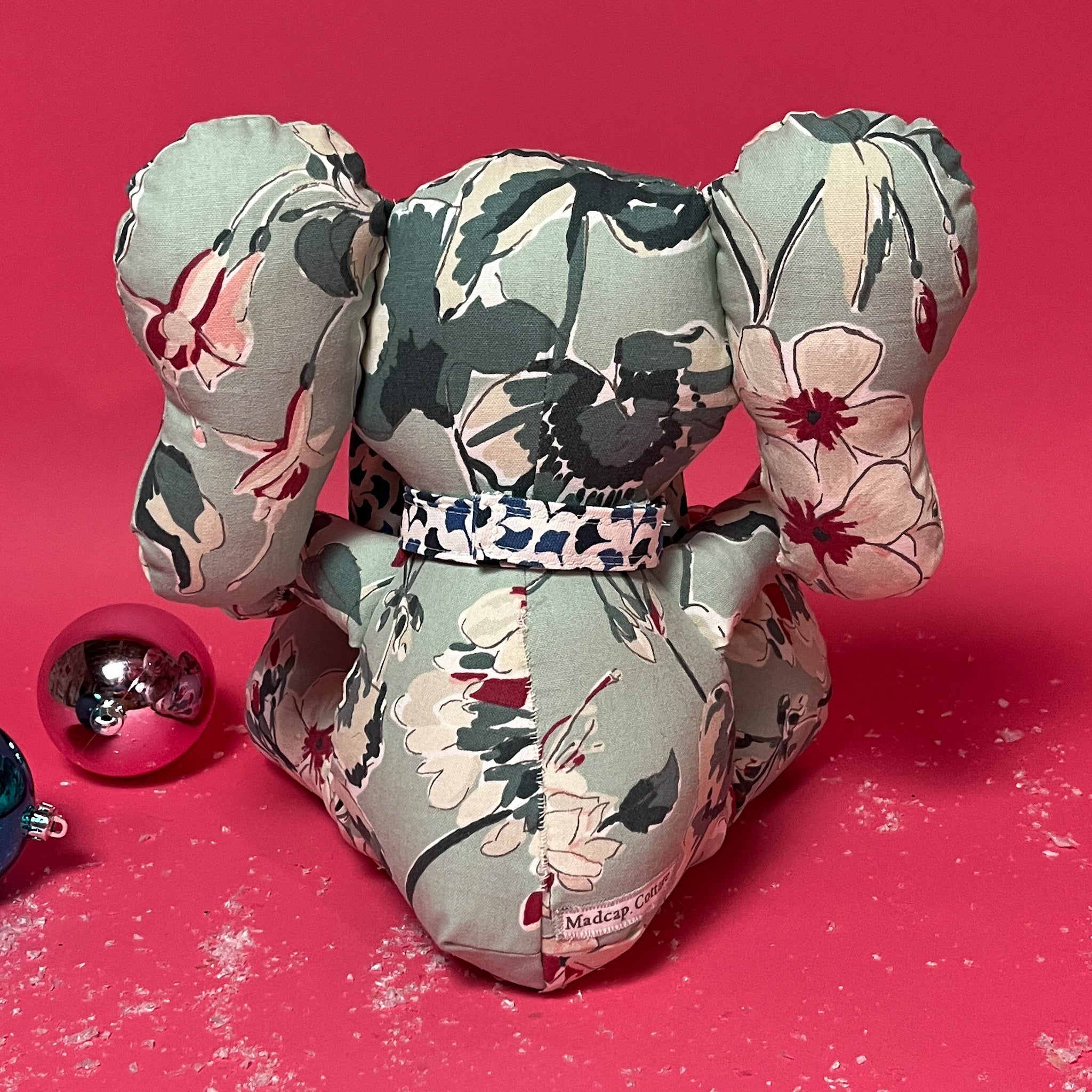 Back of a Floral patterned stuffed elephant toy with a blue and white bow in a red background.
