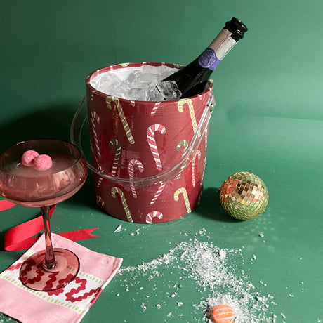 Dancing Candy Canes is a red acrylic Christmas ice bucket covered in a candy cane motif fabric filled with ice and champagne and Christmas decorations in the image foreground.