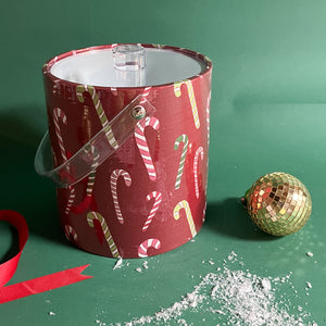 A red acrylic ice bucket covered in a candy can motif fabric with faux snow and Christmas decorations in the image foreground. 