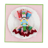 square pink paper placemats with colourful ornaments motifs in the center surrounded by a pink and green border.  A white plate is placed on top of it with a cloth and a nutcraker napkin ring