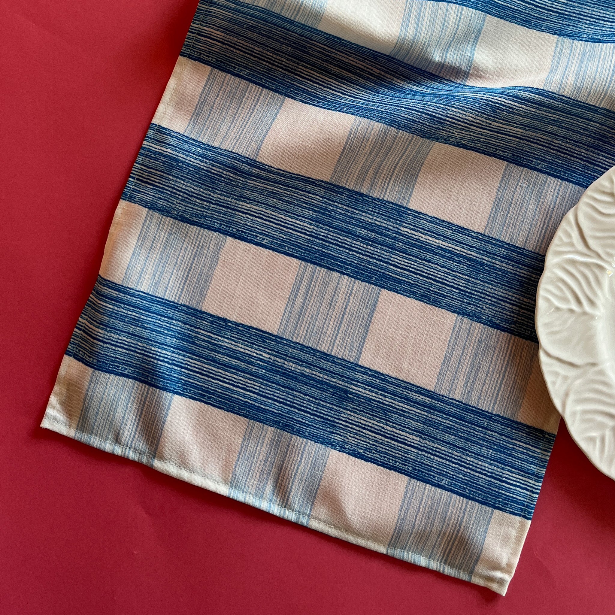 The Gin Lane cloth table runner is a gingham print in Swedish Blue shown on a red background with a white dinner plate