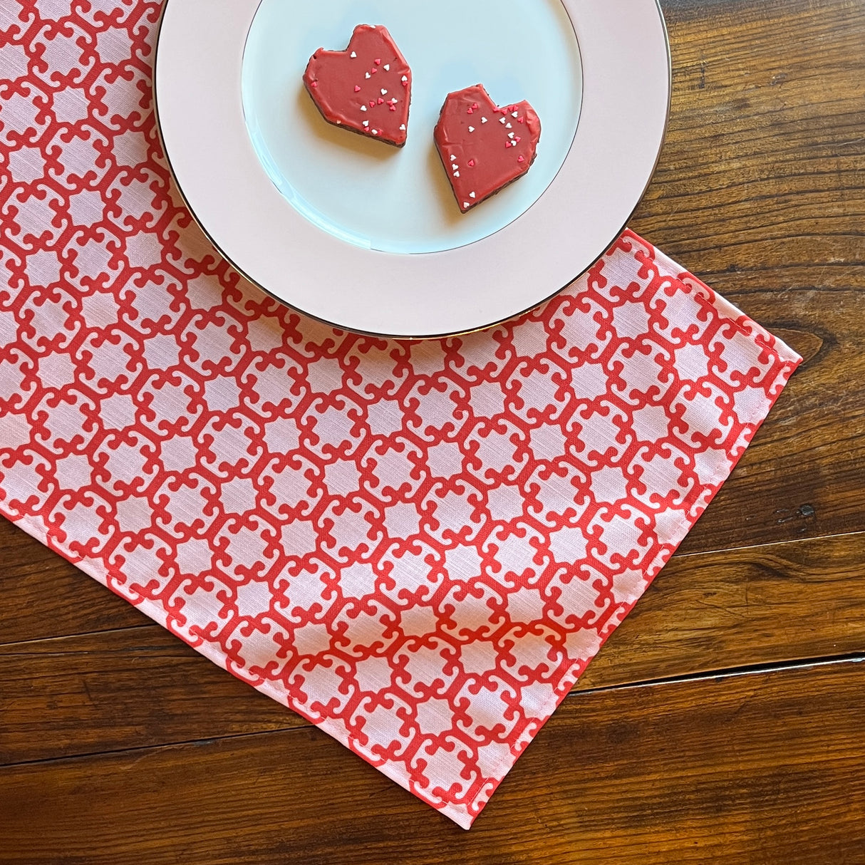 Monserrat Lacquer Red is a no-iron cloth table runner with a geometric pattern, displayed on a table with a plate of cookies on top.