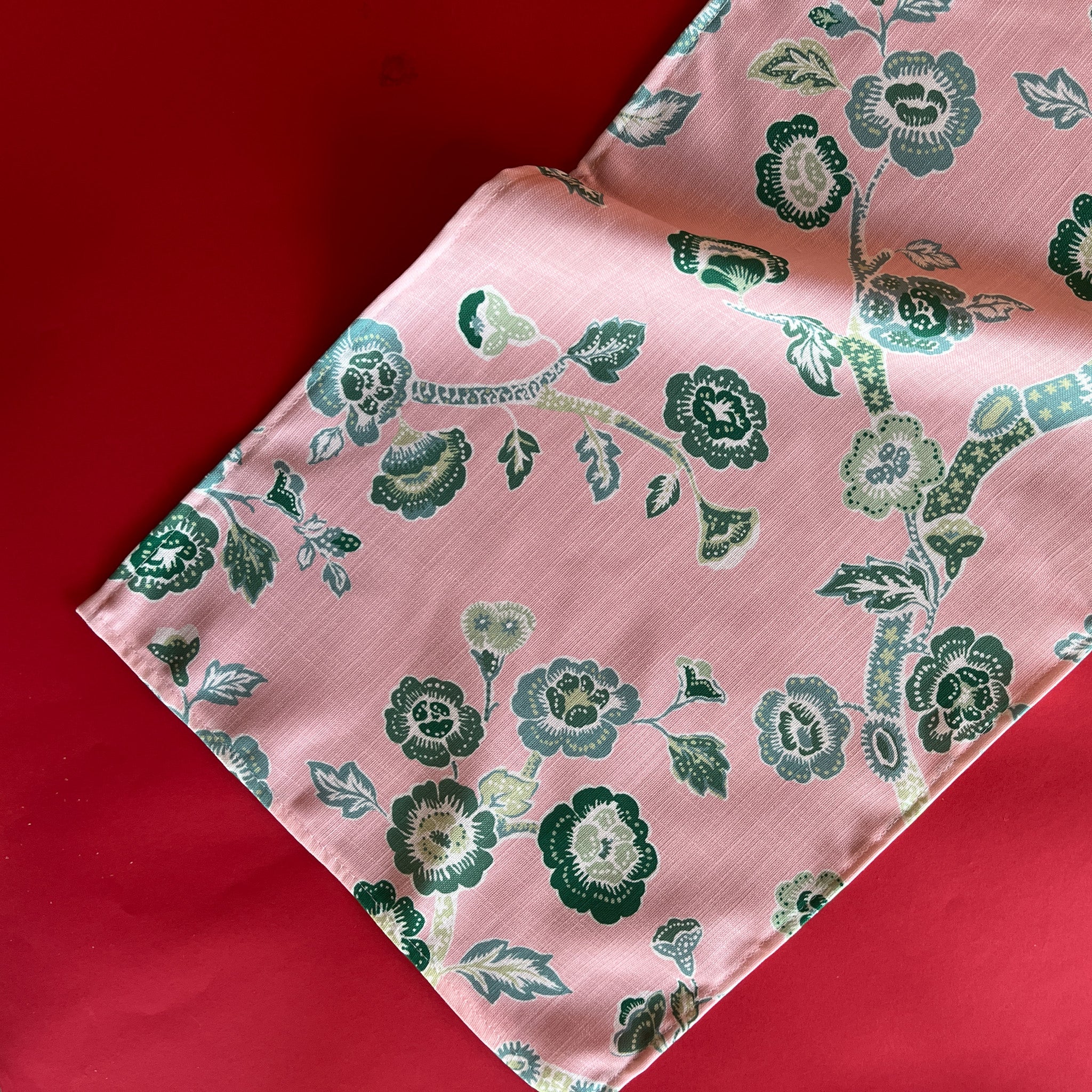 A pink Temple Garden cloth table runner with a green floral chinoiserie pattern laid out on a red surface