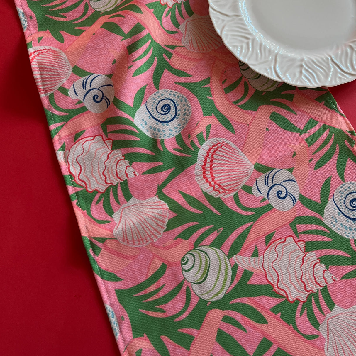 Detailed view of Shell Seekers cloth Table Runner features colorful shells, pink ribbons and green foliage on a pink background