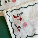 Detailed view of embroidered Christmas Jolly Santa linen cocktail napkin holding an ornament with a green scalloped border.
