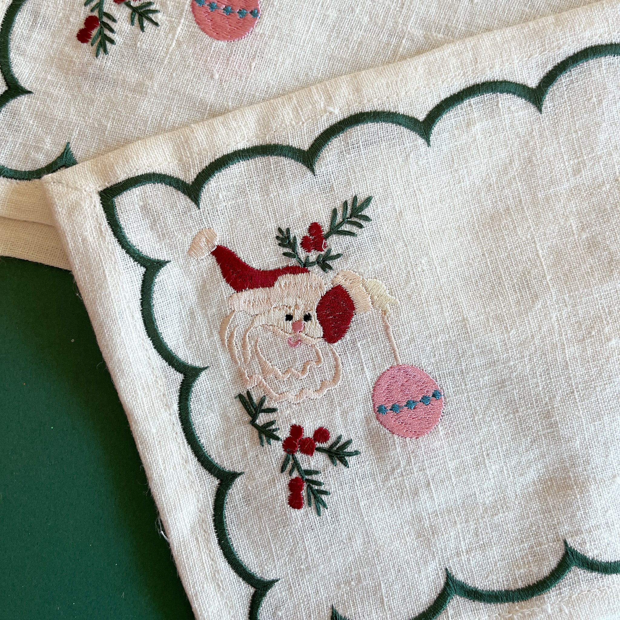 Detailed view of embroidered Christmas Jolly Santa linen cocktail napkin holding an ornament with a green scalloped border.