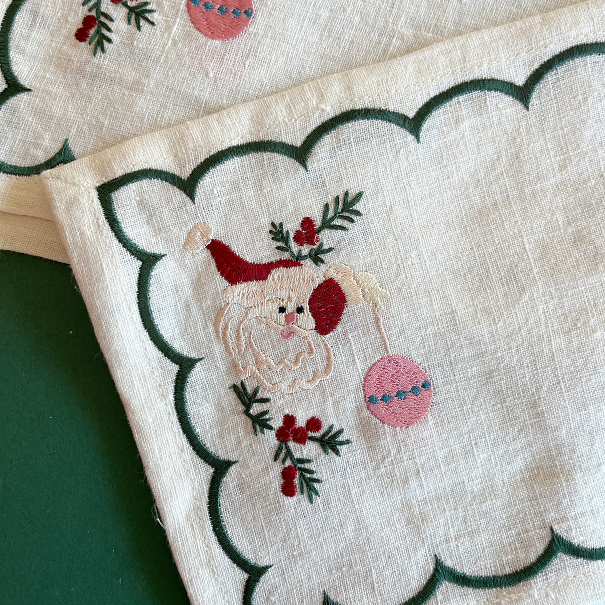 Detailed view of embroidered Christmas Jolly Santa linen cocktail napkin holding an ornament with a green scalloped border.