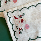 Detailed view of embroidered Christmas Jolly Santa linen cocktail napkin holding an ornament with a green scalloped border.