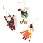 Three felt mouse ornaments dressed for winter—one in a red coat and plaid hat tossing a snowball, one in an orange coat with a yellow scarf holding a snowball, and one in a green sweater holding red berries—lying in white surface.