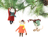 Three felt mouse ornaments dressed for winter—one in a red coat and plaid hat tossing a snowball, one in an orange coat with a yellow scarf holding a snowball, and one in a green sweater holding red berries—shown hanging on a tree
