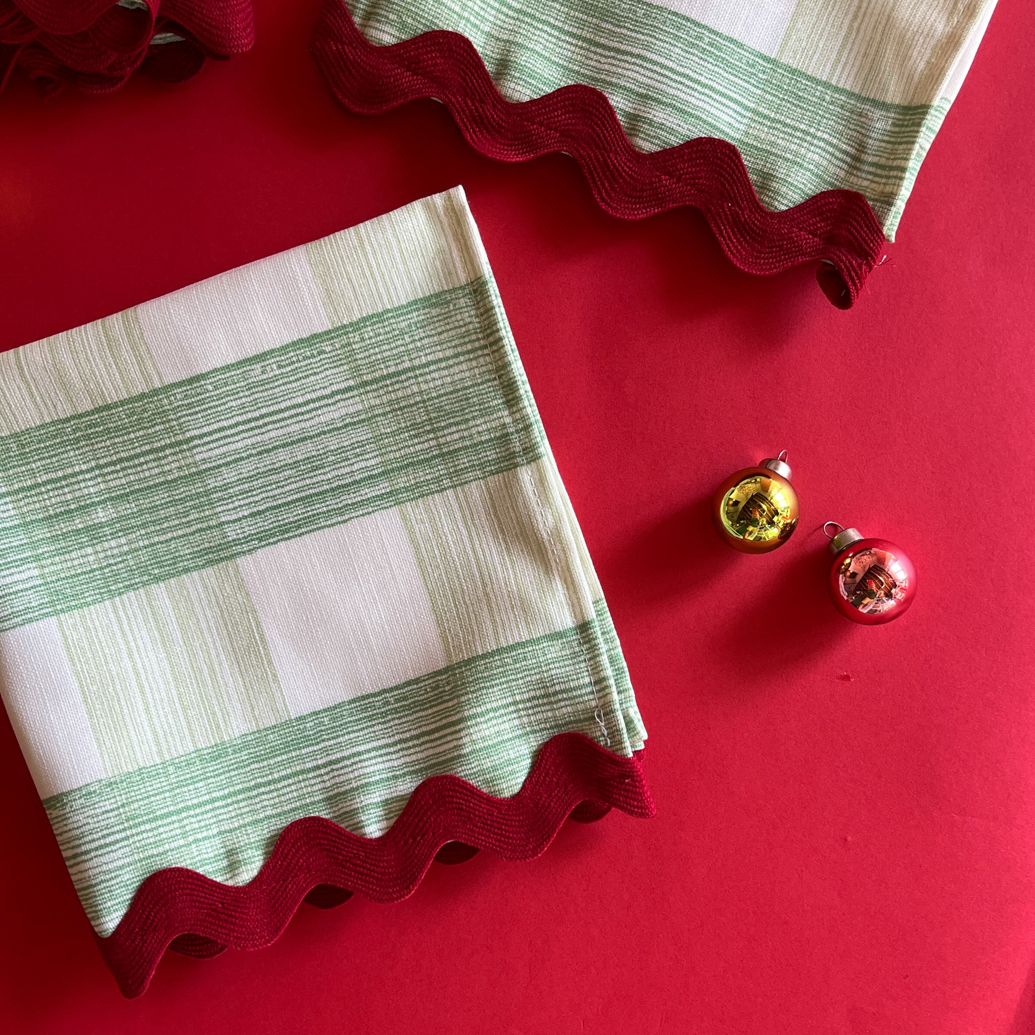 Folded Gin Lane Celery Green Gingham Cloth Dinner Napkin on a red background with two glass ornaments accenting the napkin.