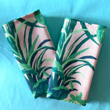 A pair of folded napkins in Jungle Road print in greens and pink on a turquoise background.