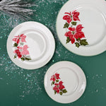 Three white dinner plates with red poinsettia floral design arranged on a green background with snow-like accents.