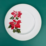 White dinner plate with a red poinsettia floral design on the left side, displayed on a green background.