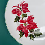 Close-up of a white dinner plate with vibrant red poinsettia flowers and green leaves on the rim.
