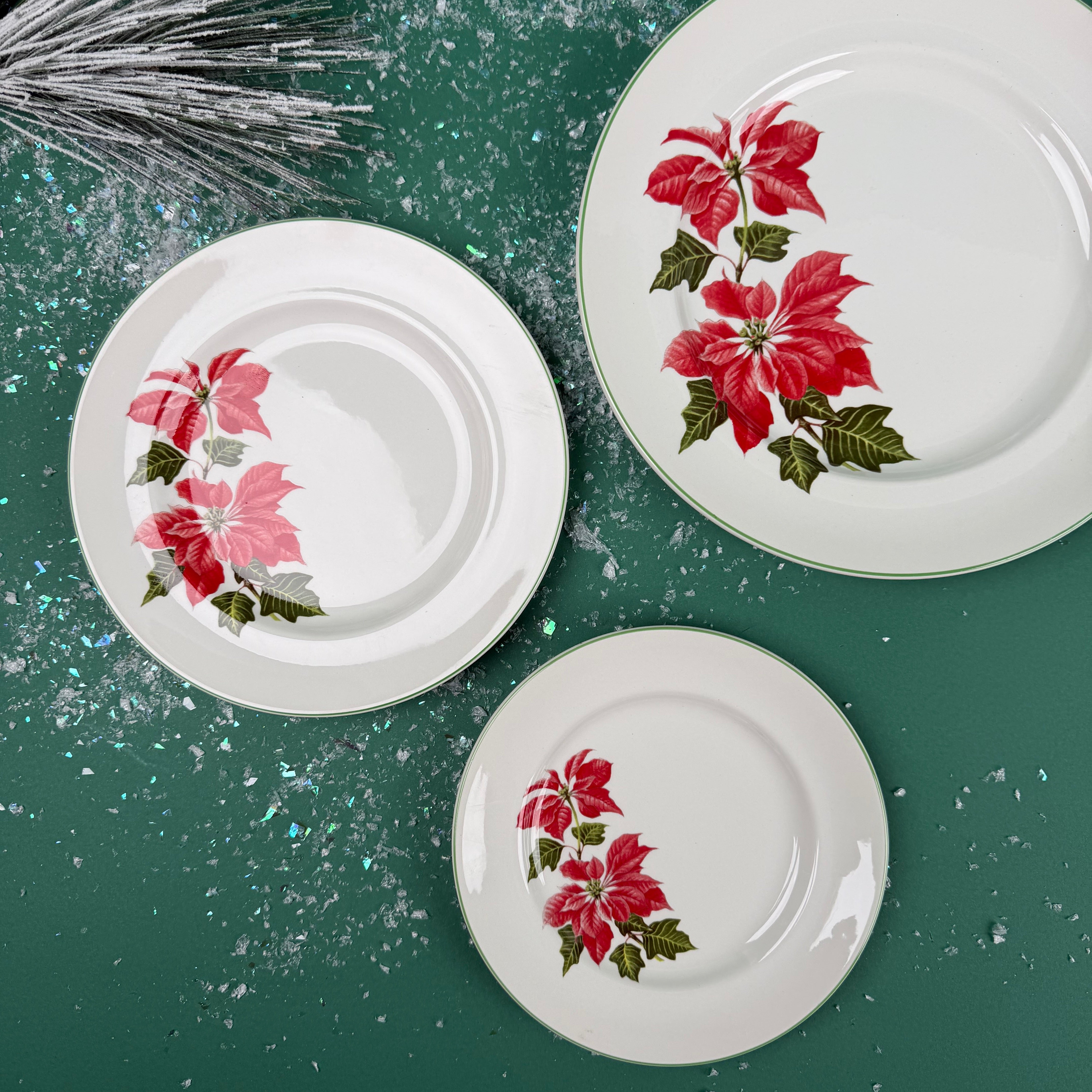 Three white dinner plates with red poinsettia floral design arranged on a green background with snow-like accents.