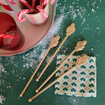 4 Gilt Metal Santa Swizzle Sticks laying on a green background with fake snow and a block print coaster
