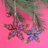 Two gilt metal star shaped Christmas ornaments decorated with colorful crystal accents hanging in a pine branch