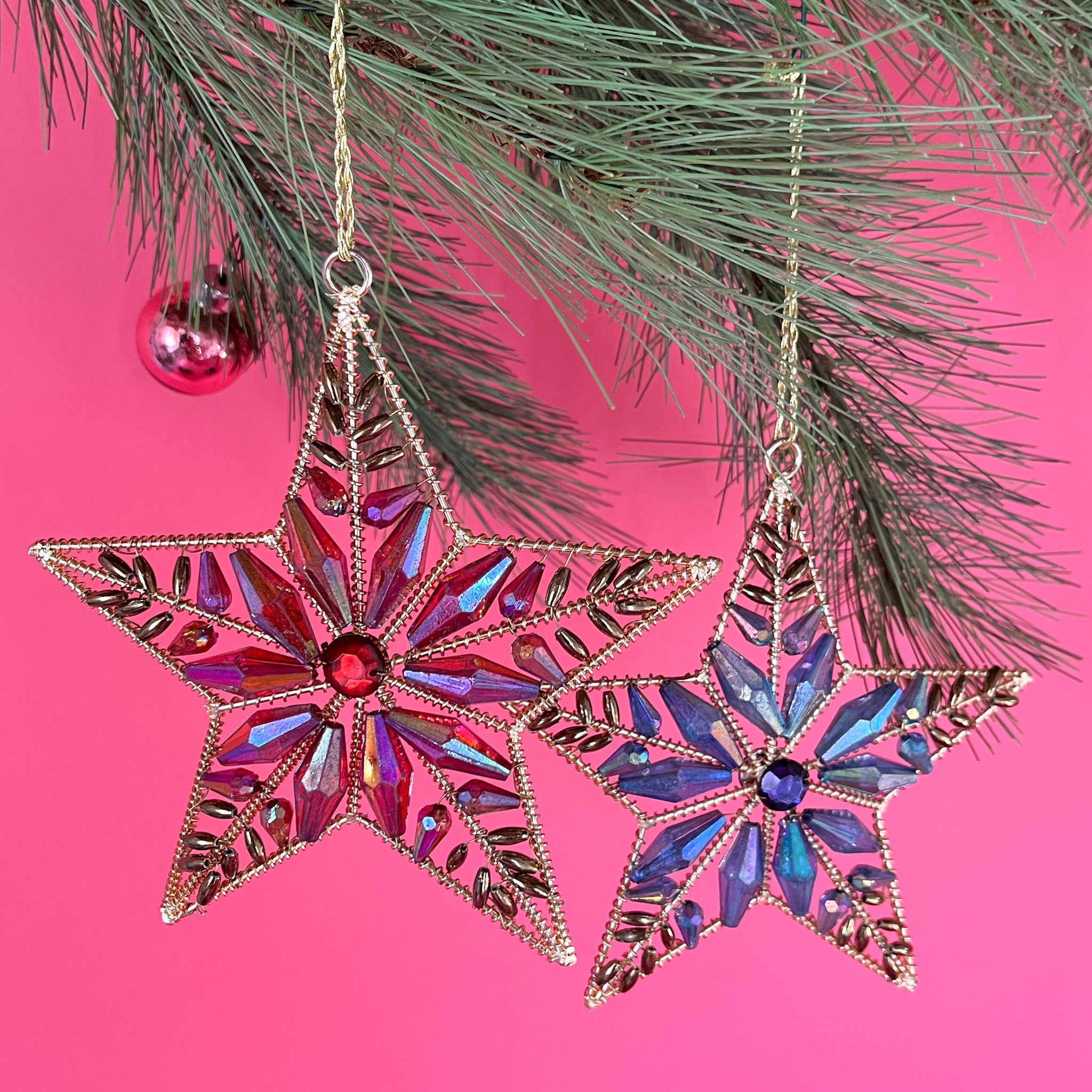 Two gilt metal star shaped Christmas ornaments decorated with colorful crystal accents hanging in a pine branch