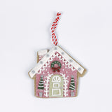 Pink gingerbread house Christmas ornament decorated with white icing details, green trees, and a wreath above the door, hanging from a red and white string.
