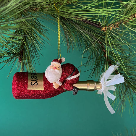 Glittery Ornament featuring santa riding a champagne bottle against a green background and topped by a pine branch. 