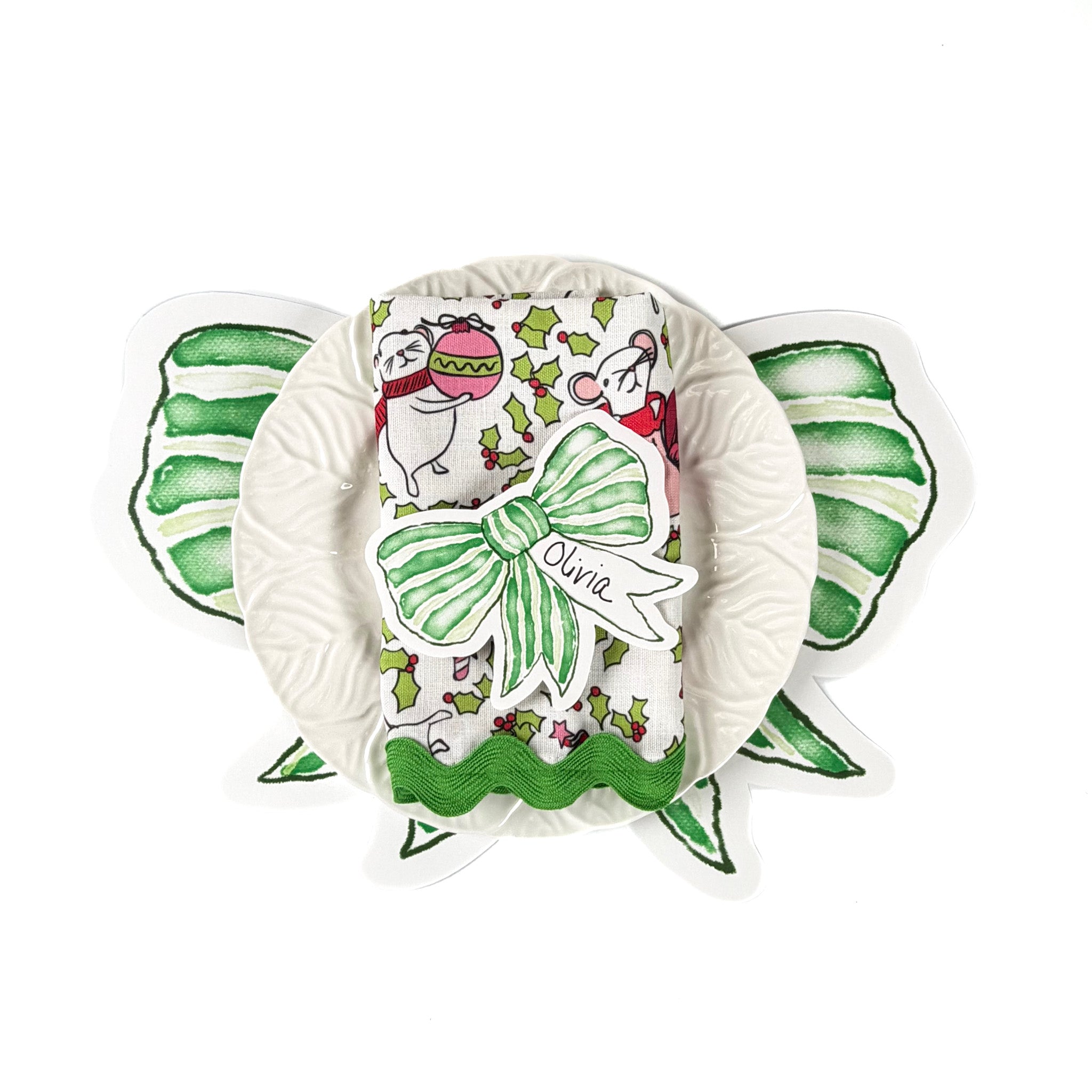 Christmas tabletop display featuring green bow–shaped paper napkins, a white ruffled plate, and a holiday napkin with festive mouse and holly pattern, arranged on a white background.